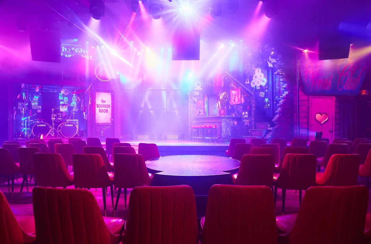 Rows of chairs sit around a stage, which is theatrically lighted with blue, white and pink spotlights.