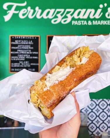 A hand holds an artichoke-and-stracciatella sandwich against a green wall with Ferrazzani's Pasta & Market written in cursive