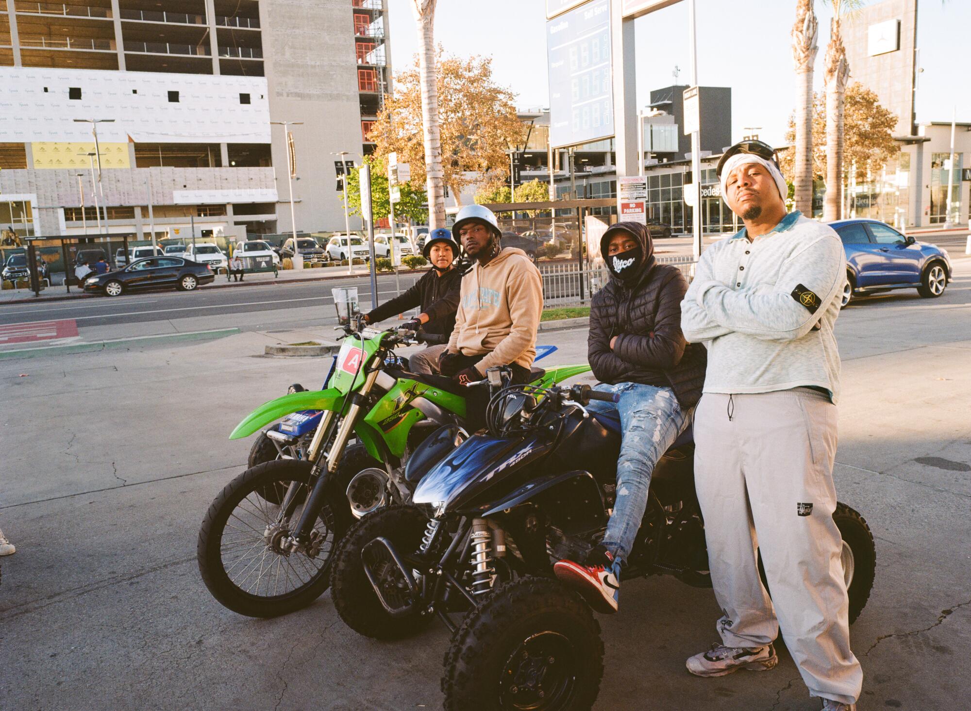 Three men sit on dirt bikes on a city street while a fourth stands next to them, arms folded.