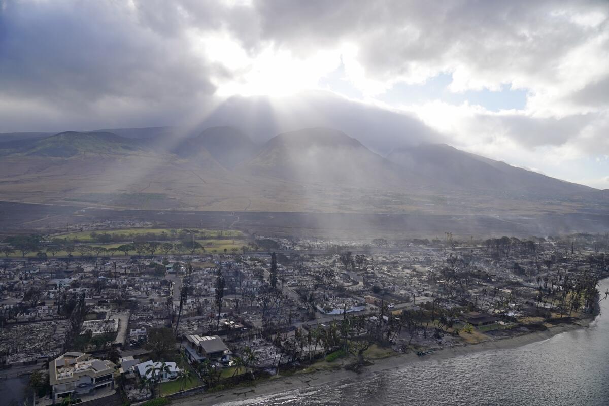 An aerial view of burned buildings alongside a body of water with the sun's rays coming through clouds above.