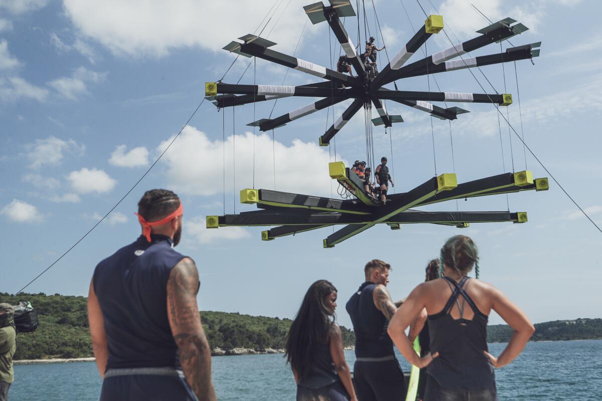 A group of reality show contestants look up at two competition platforms suspended over water.