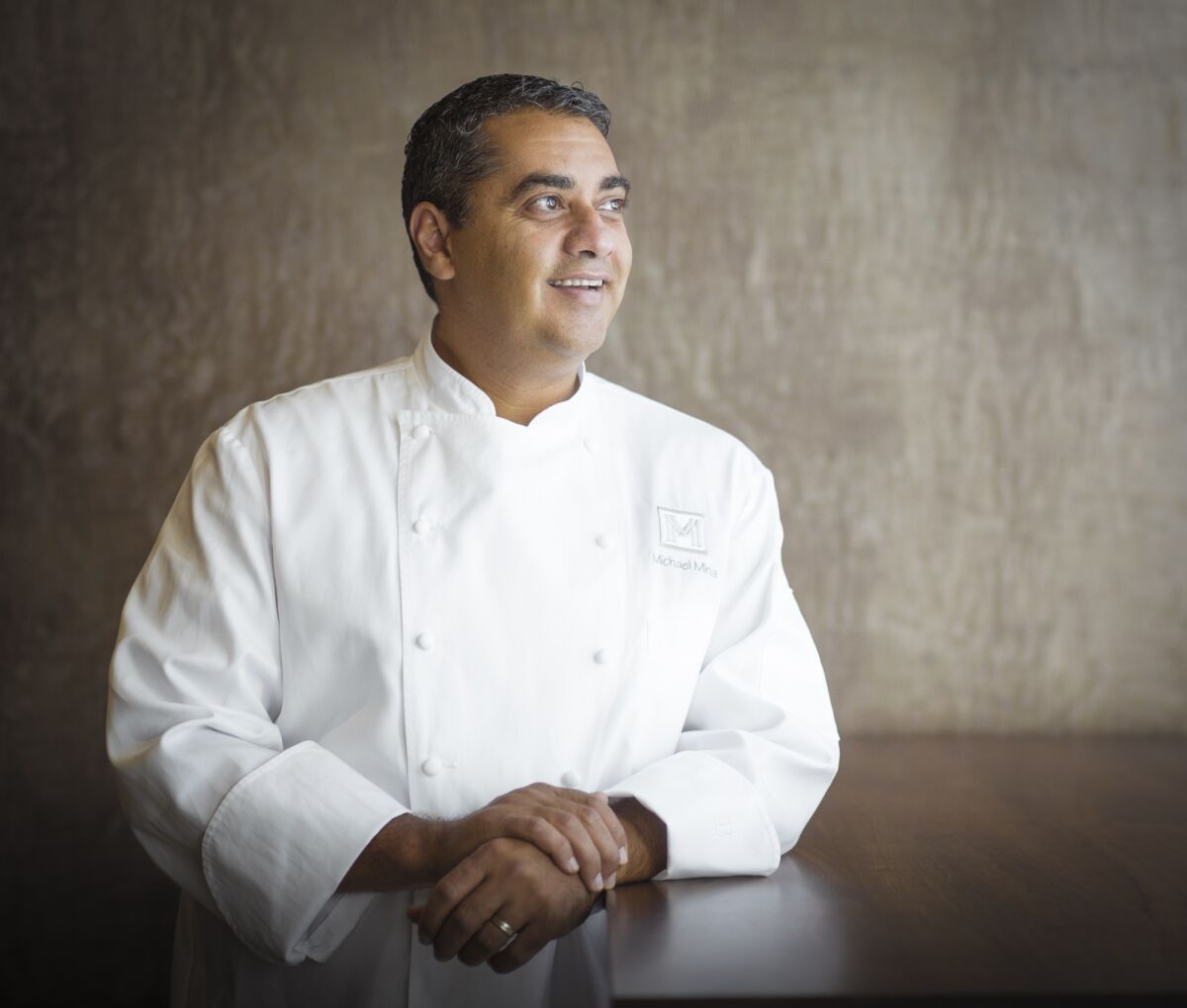 A smiling man in chef's whites leans on a counter.