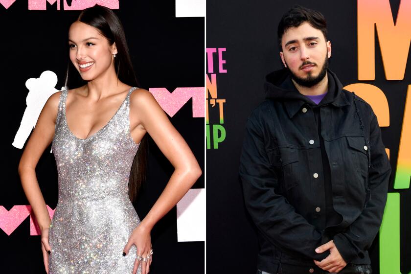 Olivia Rodrigo smiling and posing in a long silver gown. A man with dark hair and facial hair in a black outfit