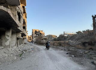 Two people on a motorcycle pass destroyed buildings, with rubble on the ground