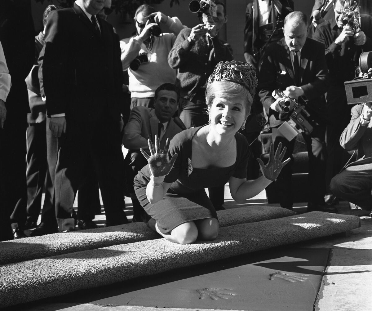 Debbie Reynolds holds up dirty hands as she kneels on a carpeted step. She wears a sheath dress and pillbox hat.