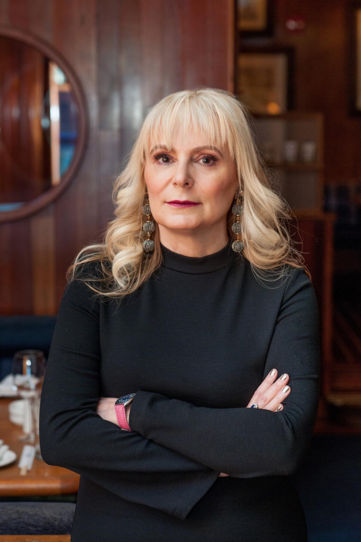 Woman with blond hair and her arms crossed wearing black long-sleeved shirt stands in a wood-paneled room 