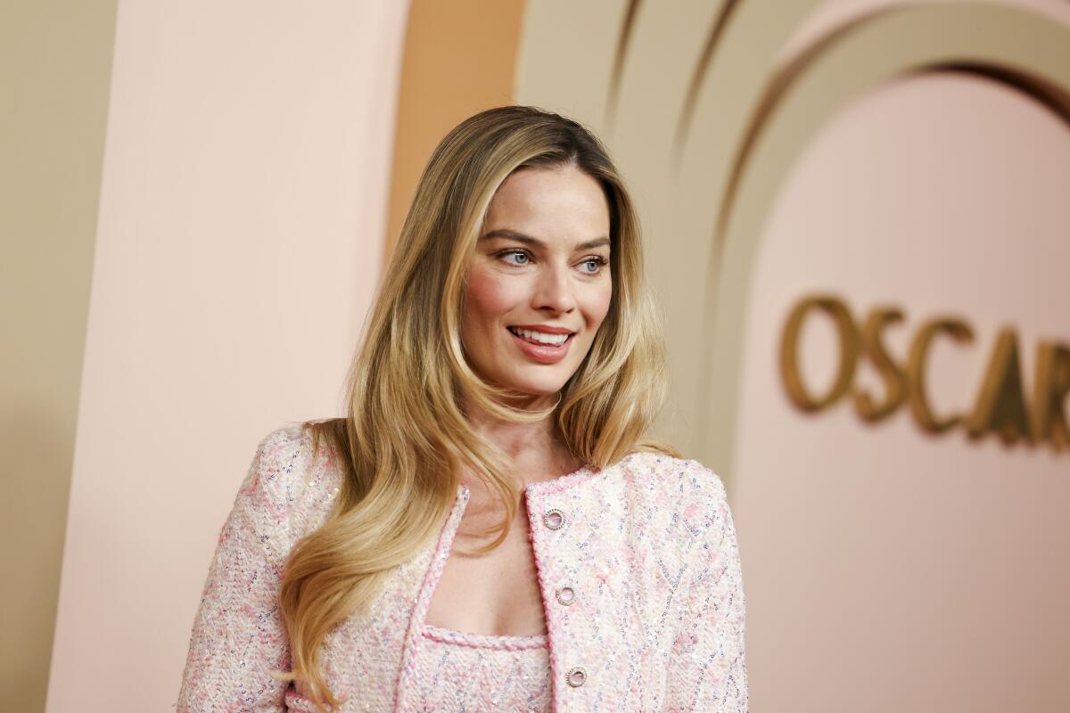 Margot Robbie wearing a pink jacket against a beige and gold backdrop