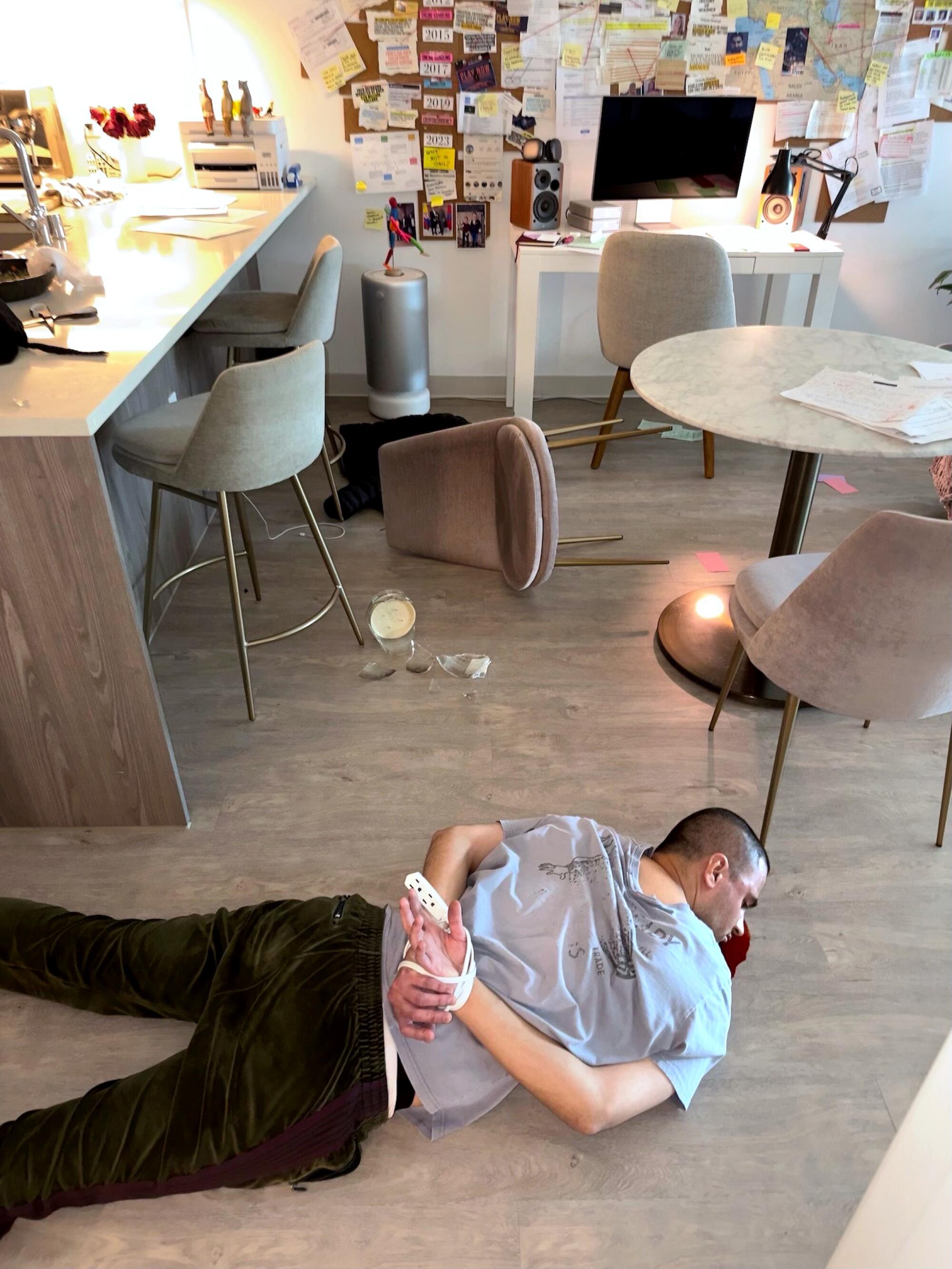 A man lays on a floor with his hands bound behind his back with an electrical cord and red liquid under his head.