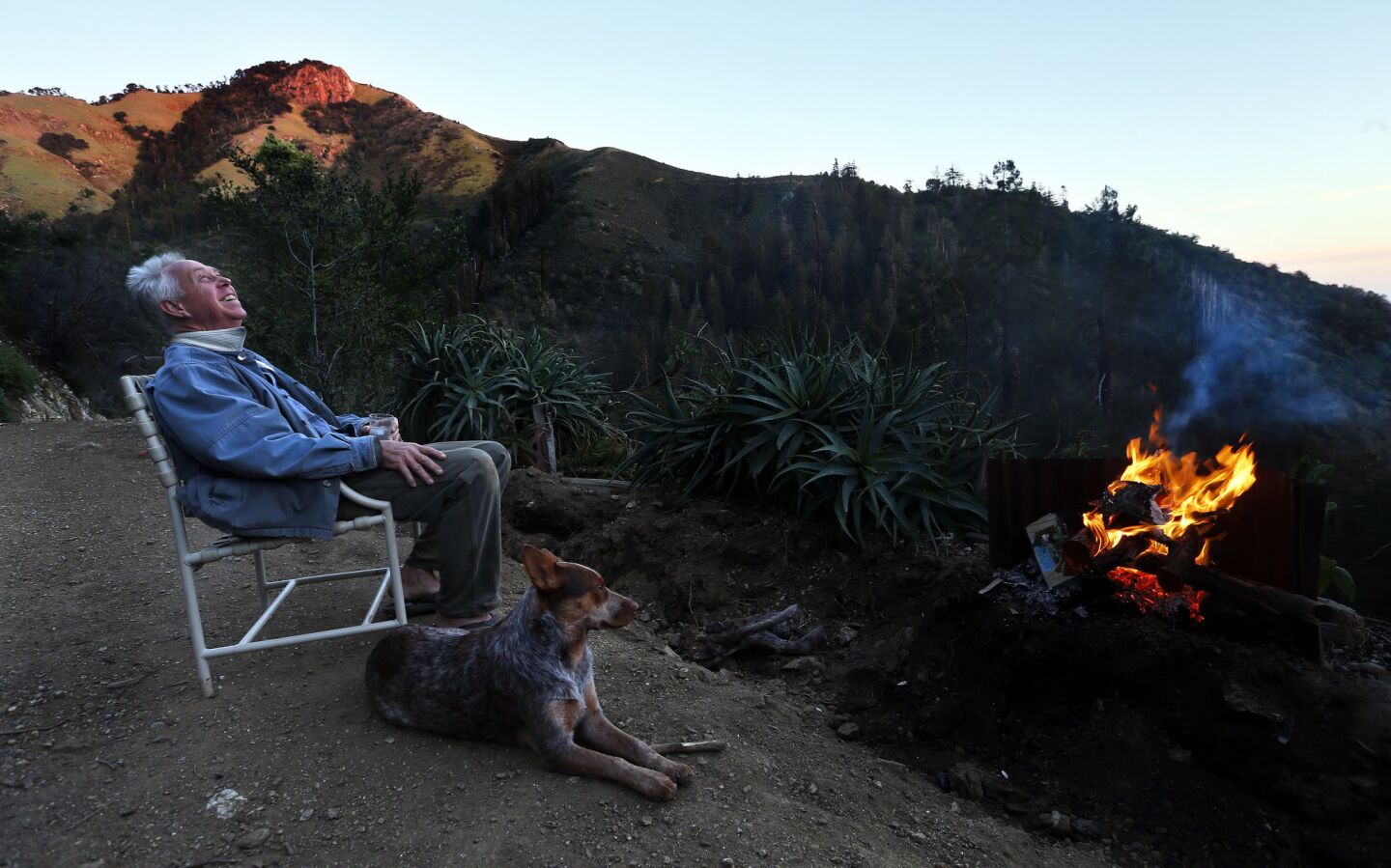 Richard Wangoe, with his dog, Zoie, an Australian heeler at his side, enjoys the warmth of a fire as he takes in the sunset from the top of his 54-acre property above Hot Springs Creek.