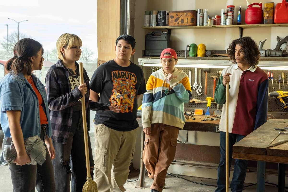 Five teenagers stand in a room holding brooms and look at one another.