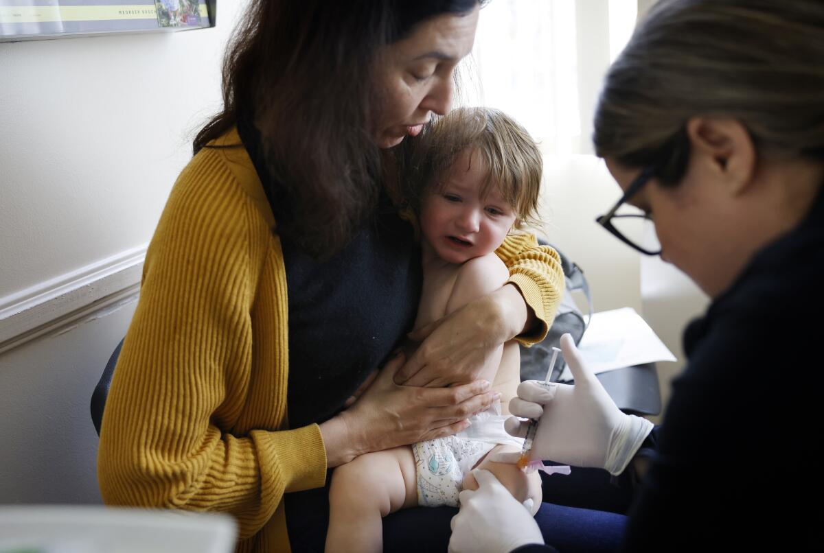 LADERA RANCH-CA-FEBRUARY 28, 2024: Karla Benzl, of Mission Viejo, center, holds her 15-month-old son Marcus while he gets his vaccinations by medical assistant Shellee Rayl at Southern Orange County Pediatric Associates in Ladera Ranch on February 28, 2024. (Christina House / Los Angeles Times)
