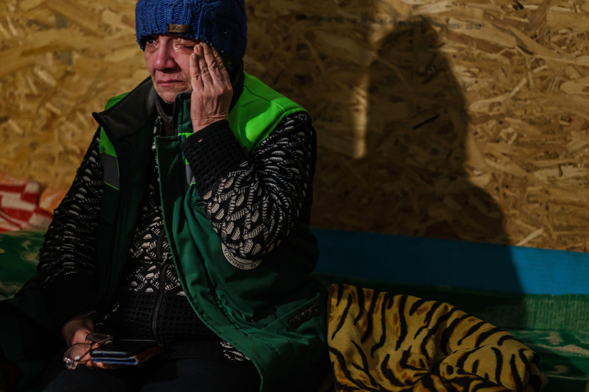 A woman wearing a blue beanie and green vest cries while seated inside a dimly lit room in a shelter.