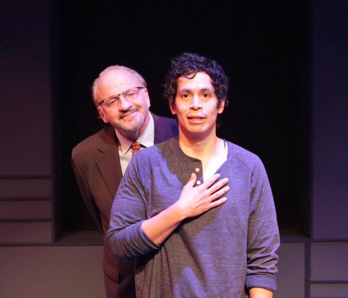 A man peers over the shoulder of a younger man who is holding his hand over his own chest.