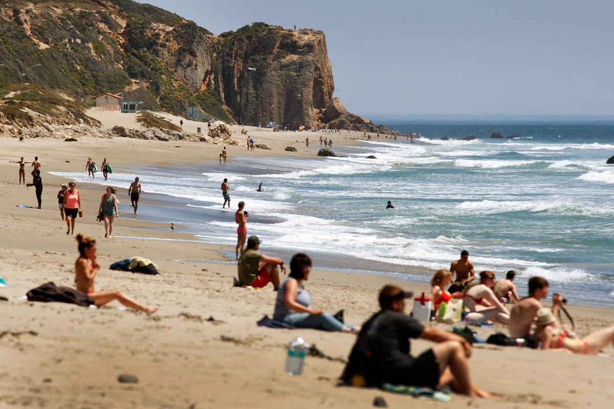 People sit on towels along the sand as waves of white foam form in the ocean.