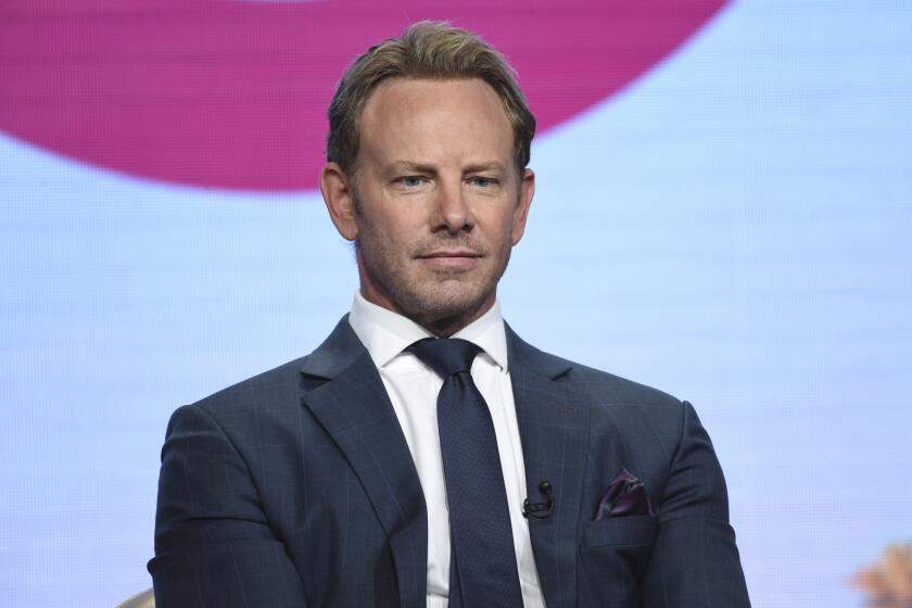 A man in a dark blue suit sitting against a large screen with white and purple imagery