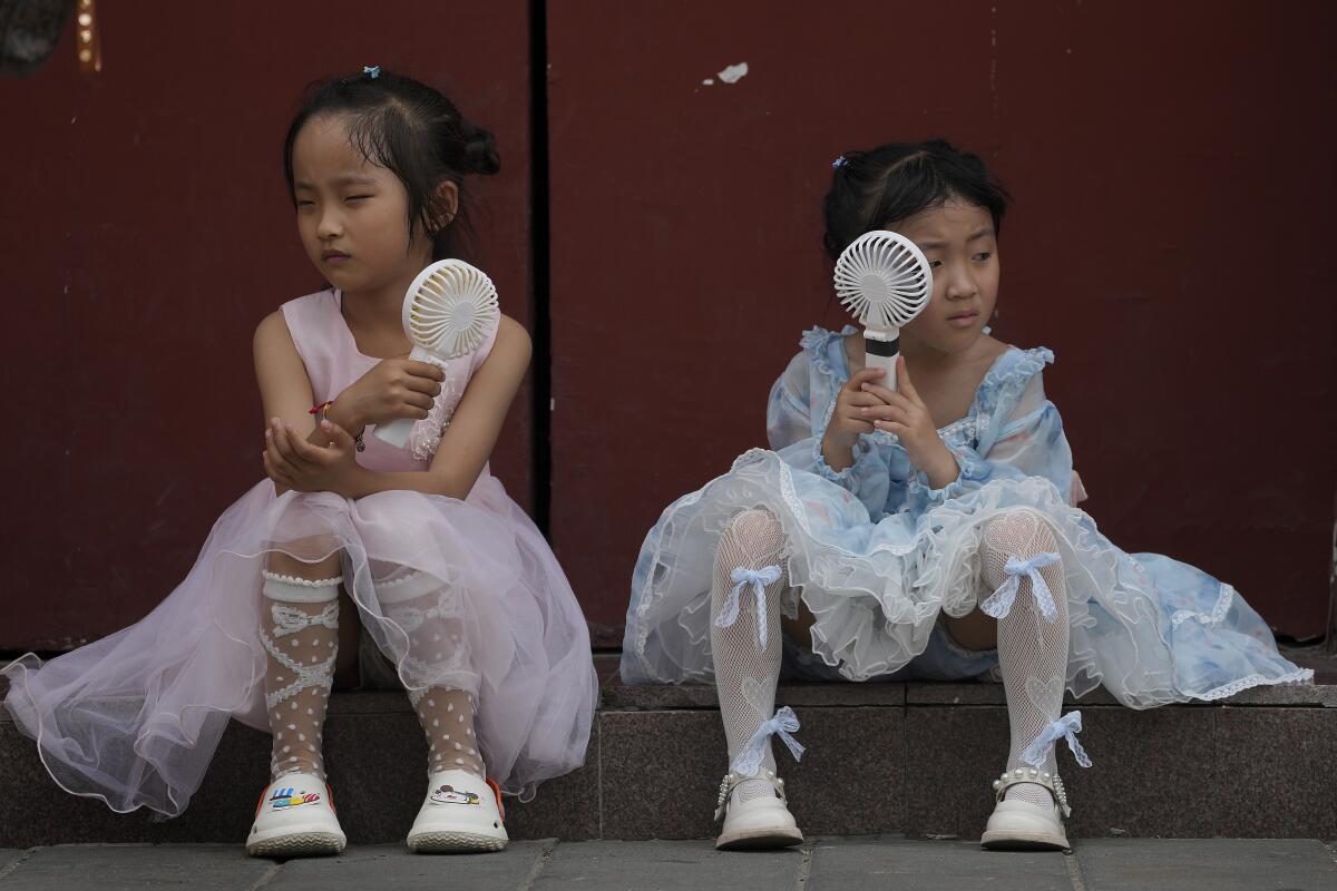 Children sit in dresses and cool themselves with handheld electric fans.