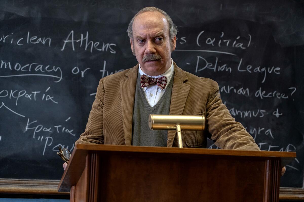 Paul Giamatti, in corduroy jacket and bowtie, stands in front of a chalkboard as the curmudgeonly teacher in "The Holdovers."