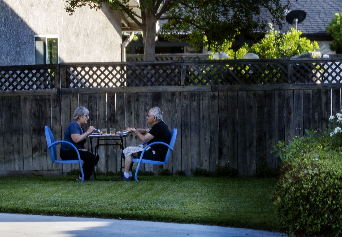 RIVERSIDE, CALIFORNIA - APRIL 27, 2020: Norm and Tracy Kahn enjoy eating dinner outside on a small cafe table sitting in blue chairs on their side yard during the coronavirus pandemic on April 27, 2020 in Riverside, California. 'During this pandemic, eating outside offers us an opportunity to change surrounding and appreciate the calmness of being outdoors among trees, scents from nature and the sounds of birds, " she said. Also adding, "Mixing up where we eat puts variety into our days and takes away the sameness of feeling trapped at home." (Gina Ferazzi/Los Angeles Times)