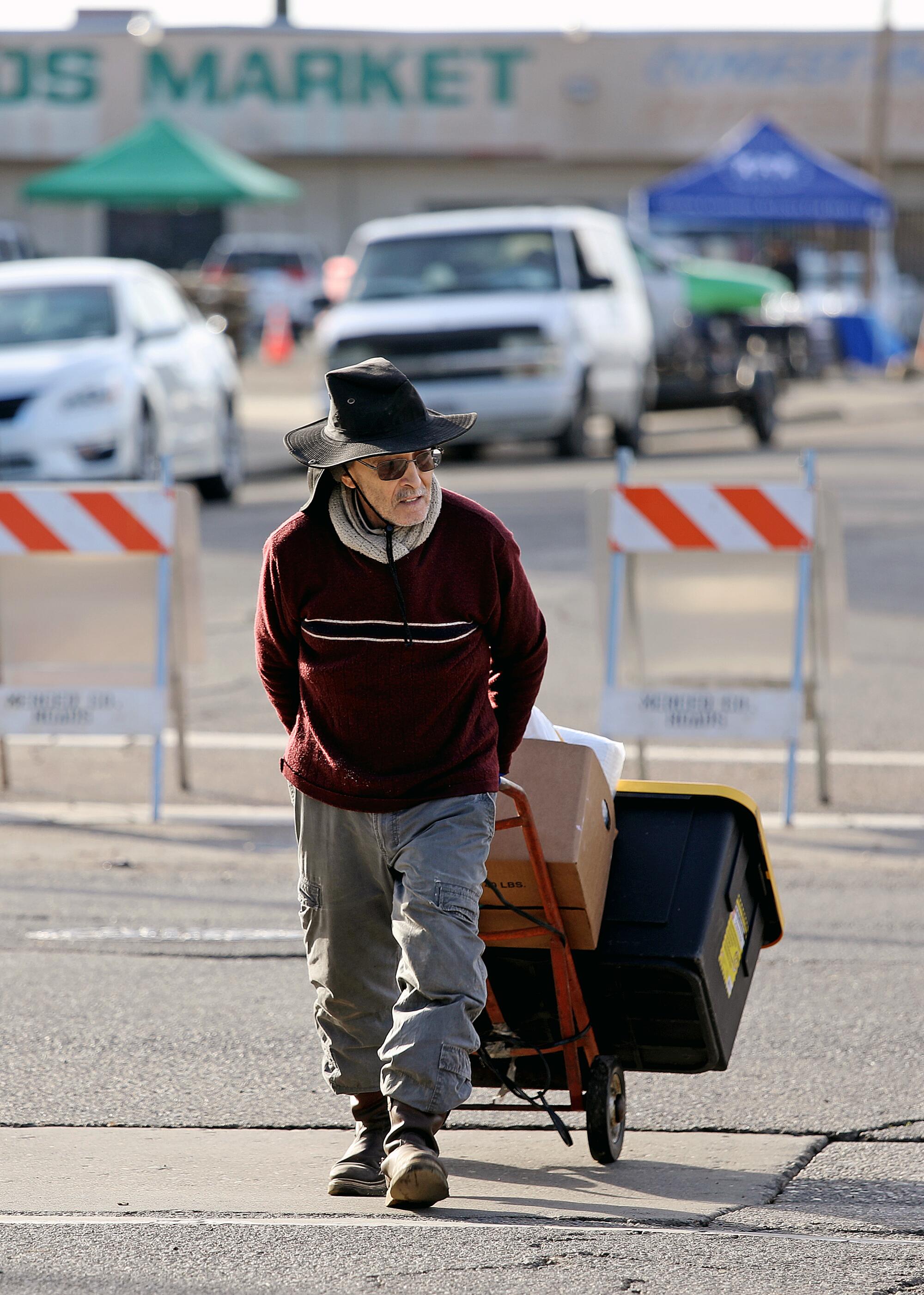A man pulls a hand cart loaded with a box and a plastic bin across a parking lot.