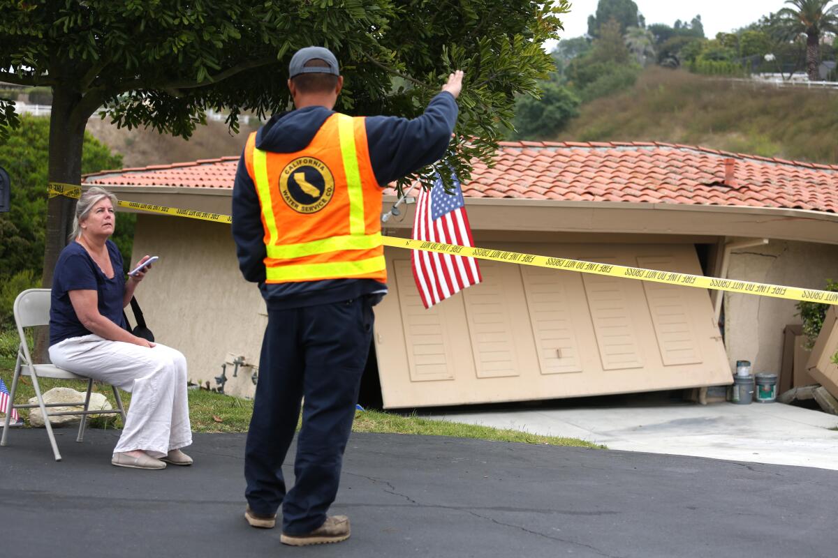 A man in a yellow and orange vest stands in the street near a damaged home and yellow tape while a woman sits in a chair.