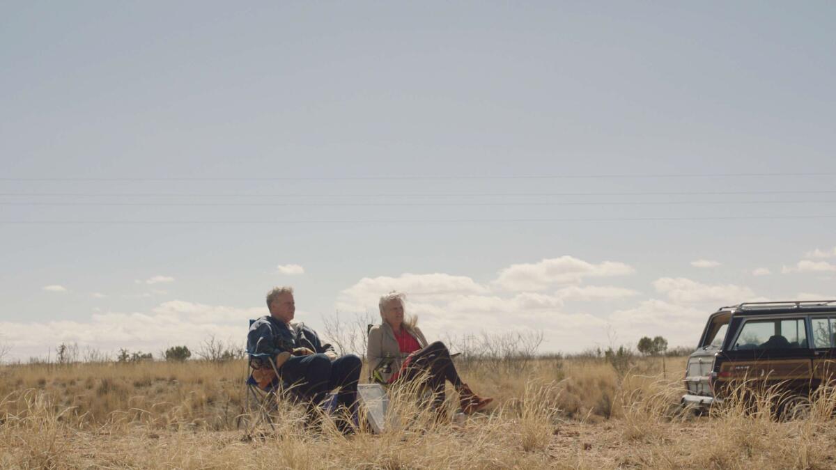 Two friends sit in camping chairs on a prairie by a station wagon.