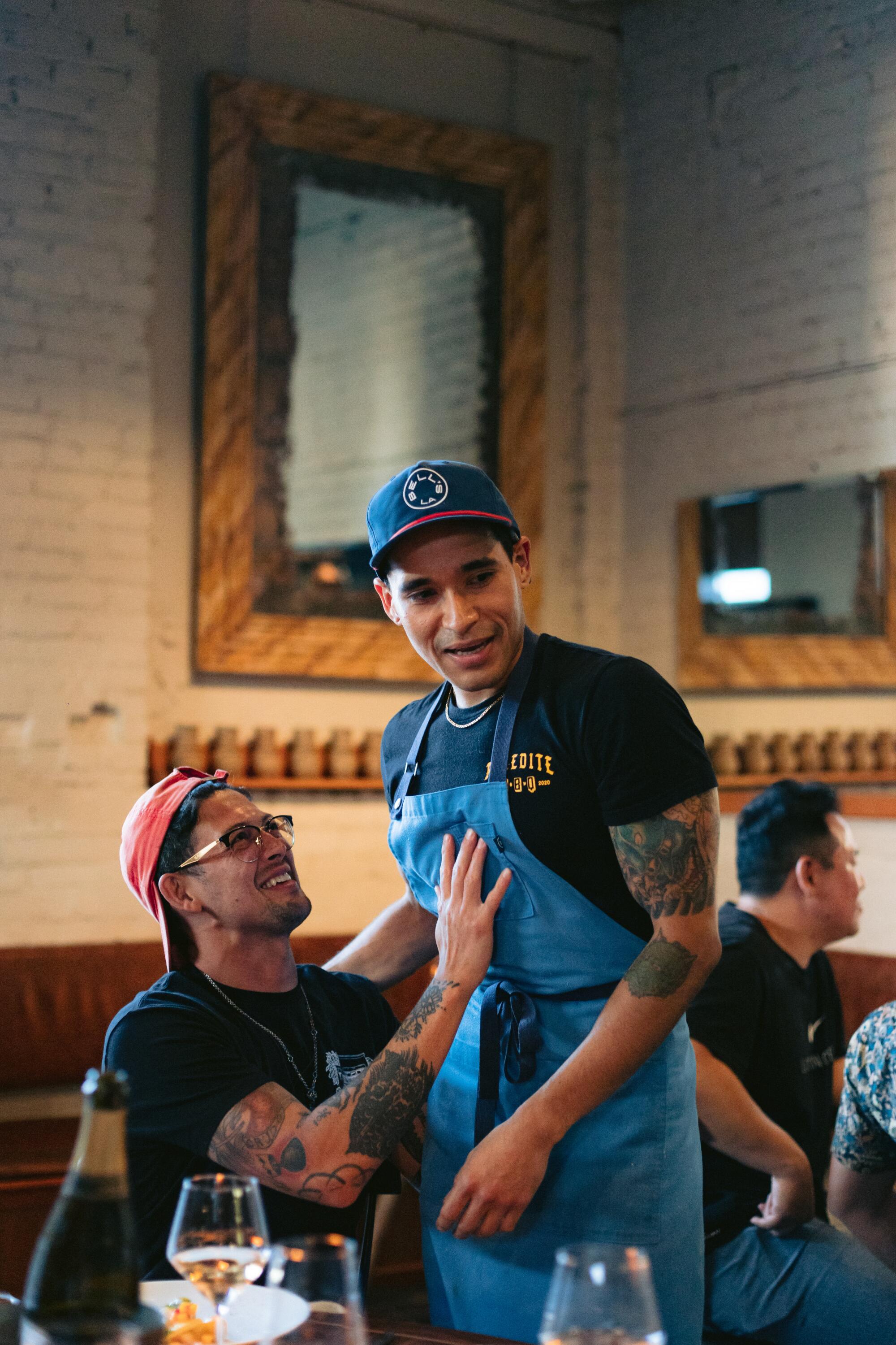 A seated man embracing a standing man in an apron at a dining table in a restaurant.