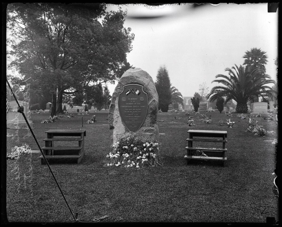 In a black-and-white photo with some damage, flowers rest at the foot of a stone monument with plaque in a cemetery. 