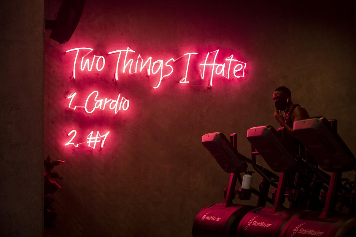 A man in a mask runs on a StairMaster machine in a dimly lit room with neon signage