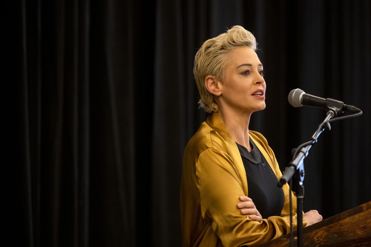 A woman with short blond hair speaks into a microphone.