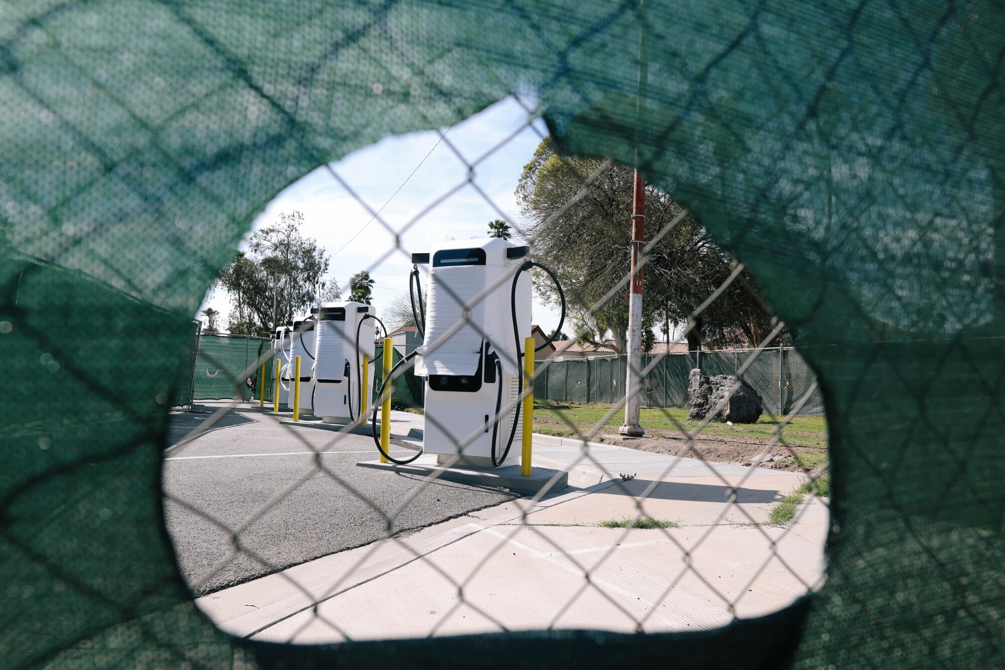 White EV charging stations seen through an opening in a mesh-covered chain link fence 
