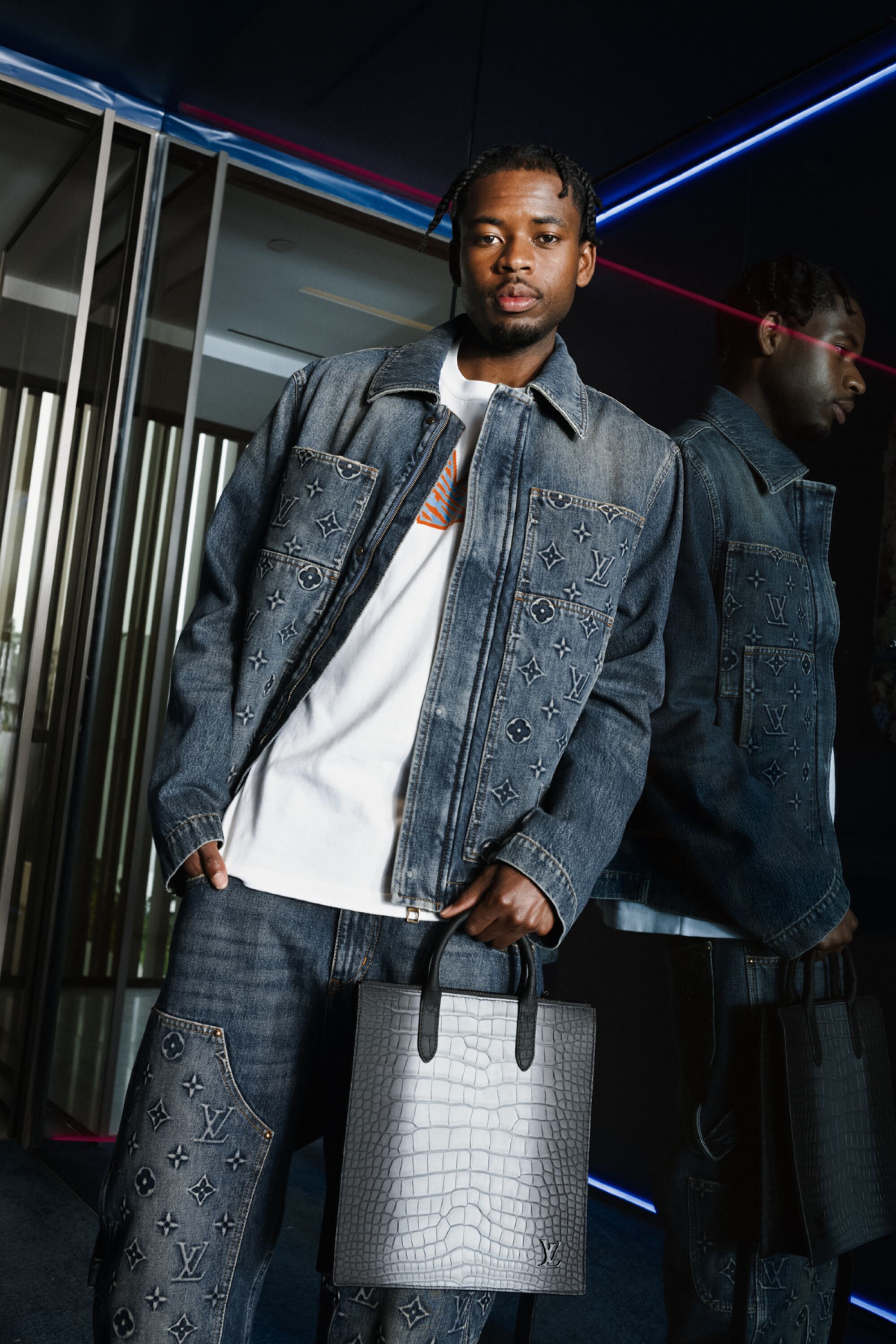 A man wearing denim leans against a mirror, holding a silver bag.