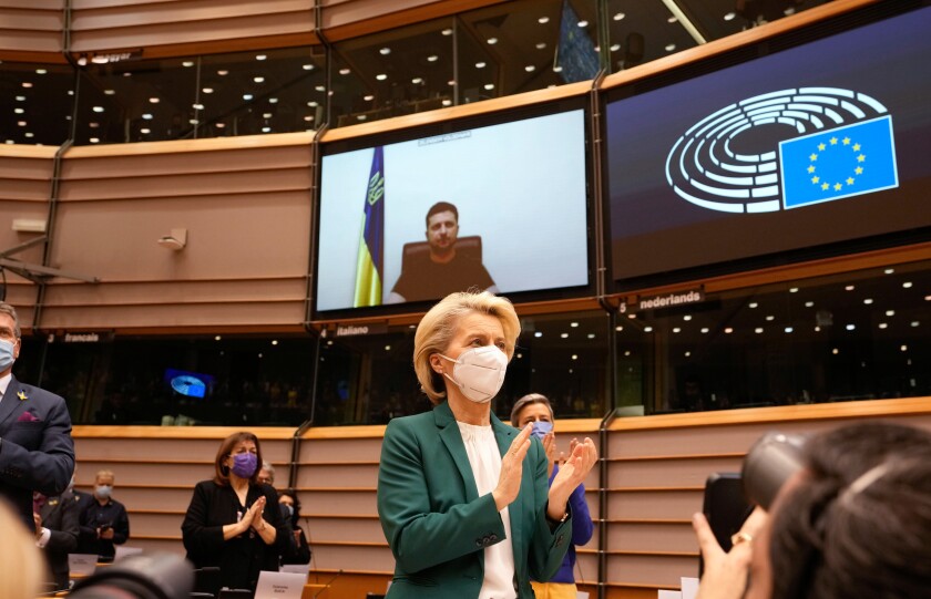 A woman in a green blazer applauds while a video screen behind her shows Ukrainian President Volodymyr Zelensky.