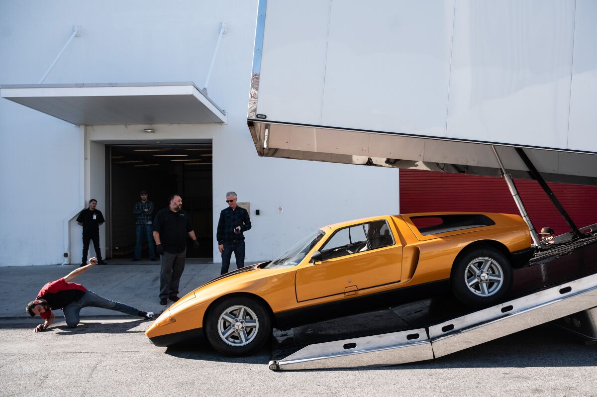 A man leans far over to watch the underside of an orange sports car as it backs into a truck