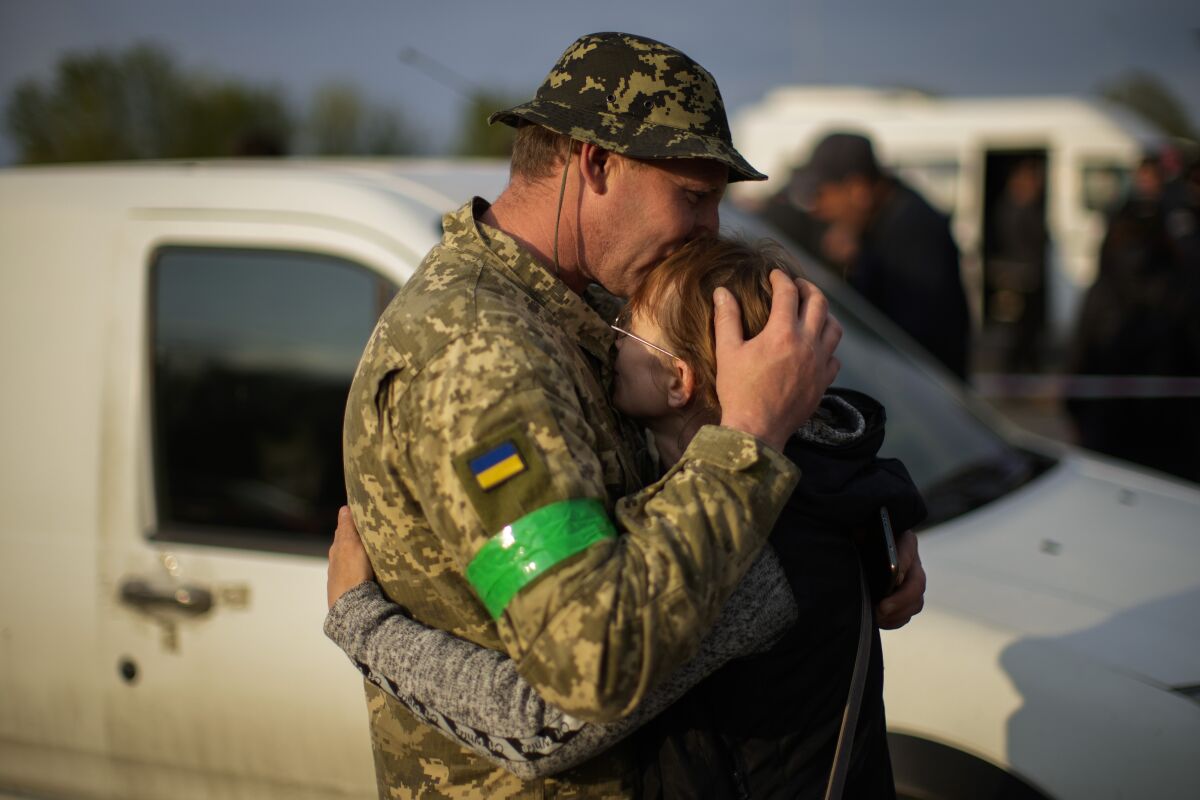 A man in fatigues, with a blue-and-yellow patch and green armband on one sleeve, embraces a woman