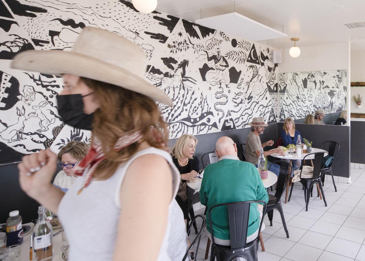 People sit and walk around in a restaurant with a black-and-white mural on the wall.