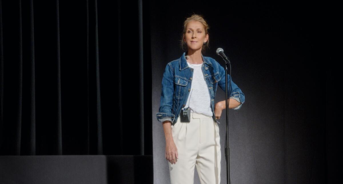 Celine Dion, wearing a denim jacket, stands in front of a mic stand.