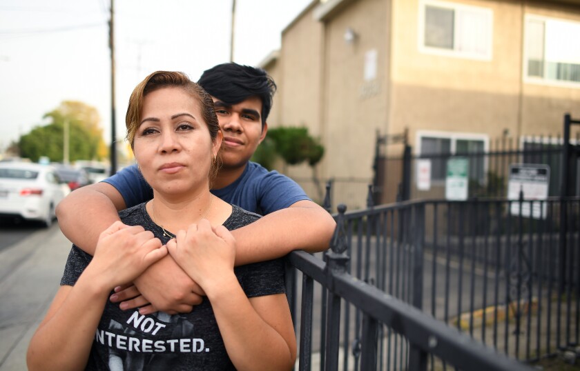 A teenage boy puts his arms around his mother as they pose for a photo in front of their apartment building