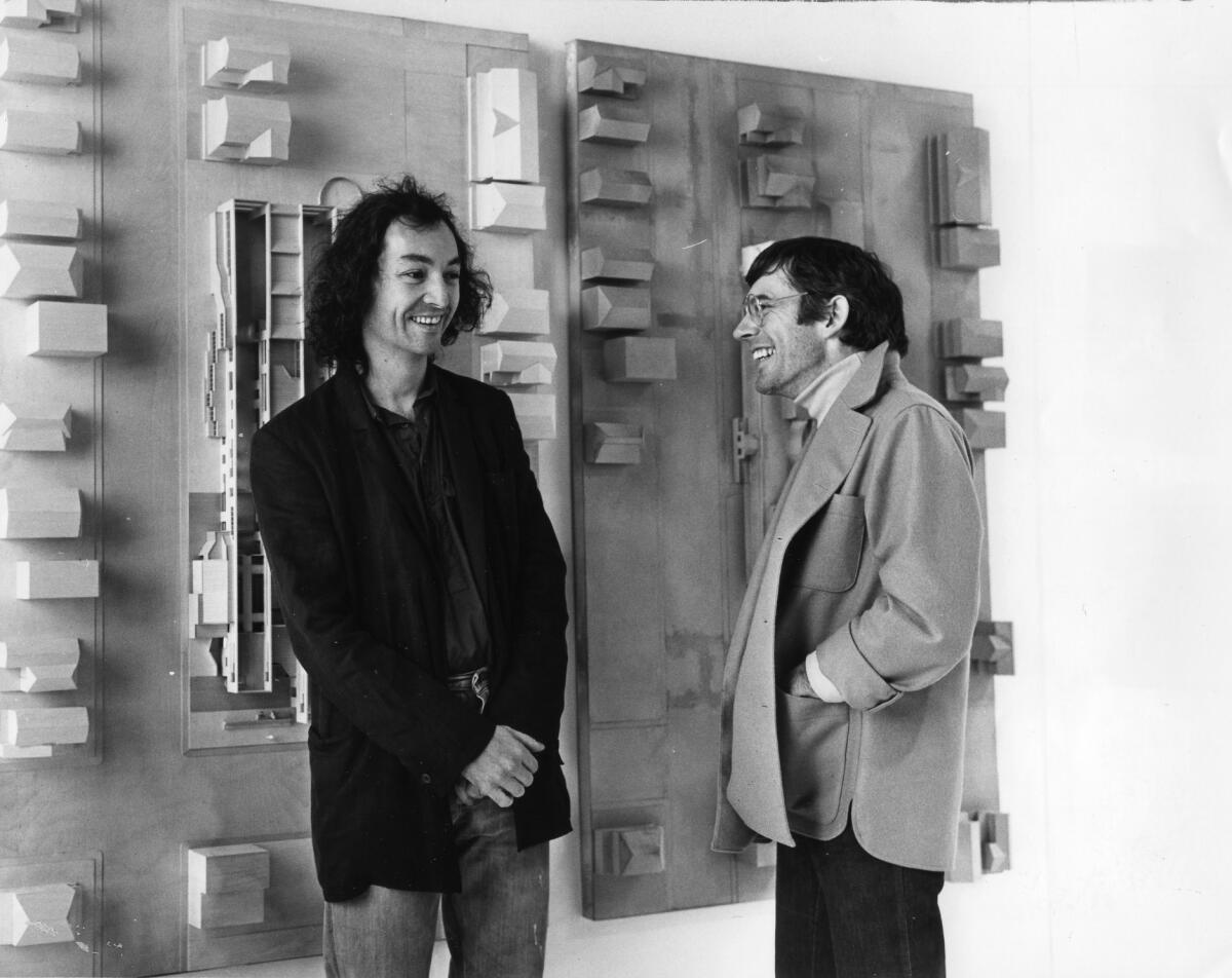 A man in a black blazer and a man in a tan jacket stand before an architectural model hanging on a wall in a vintage photo.