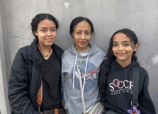 A woman with dark hair, in a gray sweatshirt, is flanked by two young girls in dark clothes