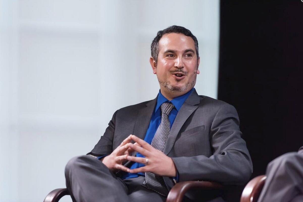 A man in a suit and tie talks and smiles as he sits in a chair on a stage. 