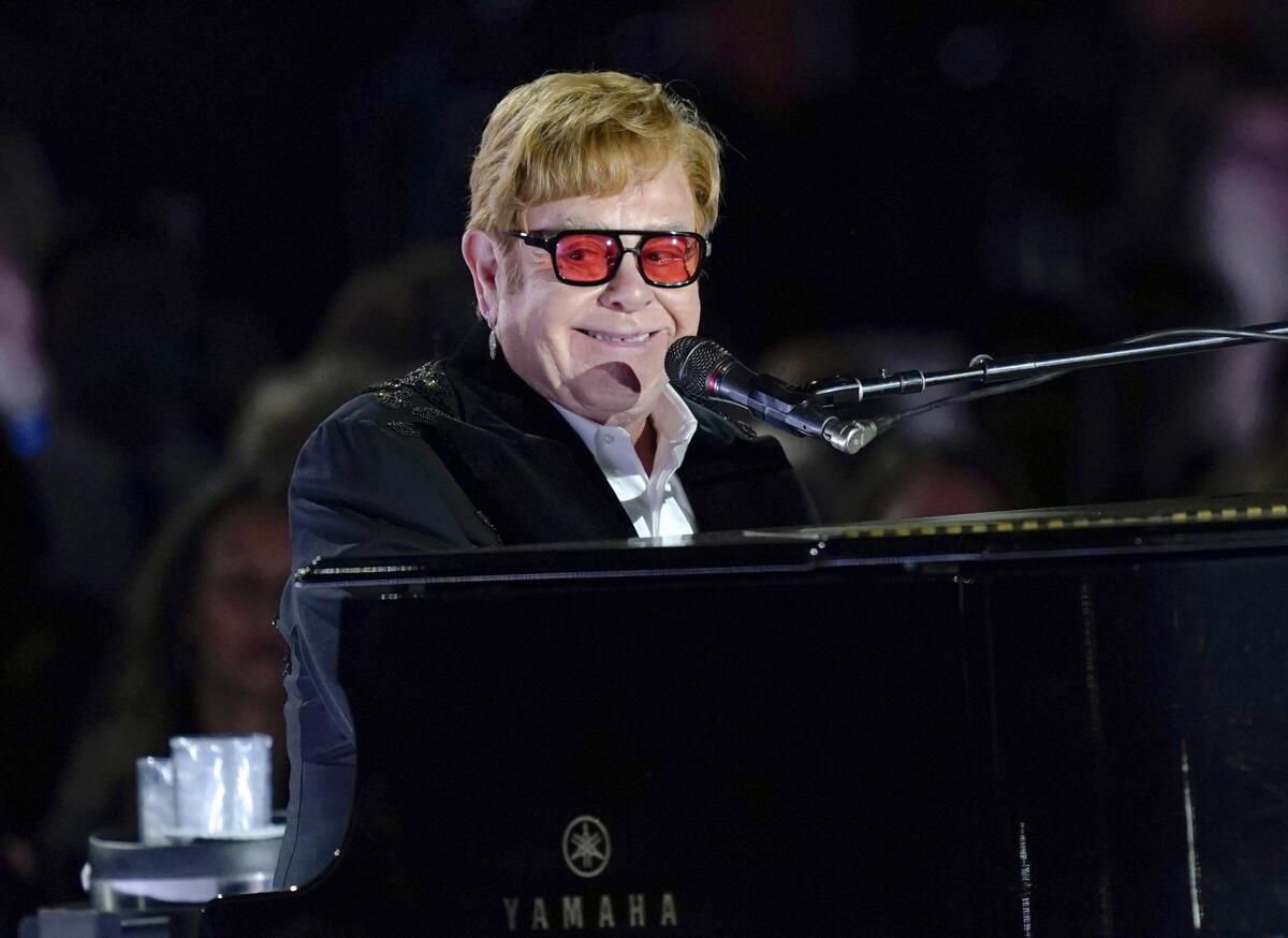 Elton John smiles while wearing red-tinted shades and playing a piano. 