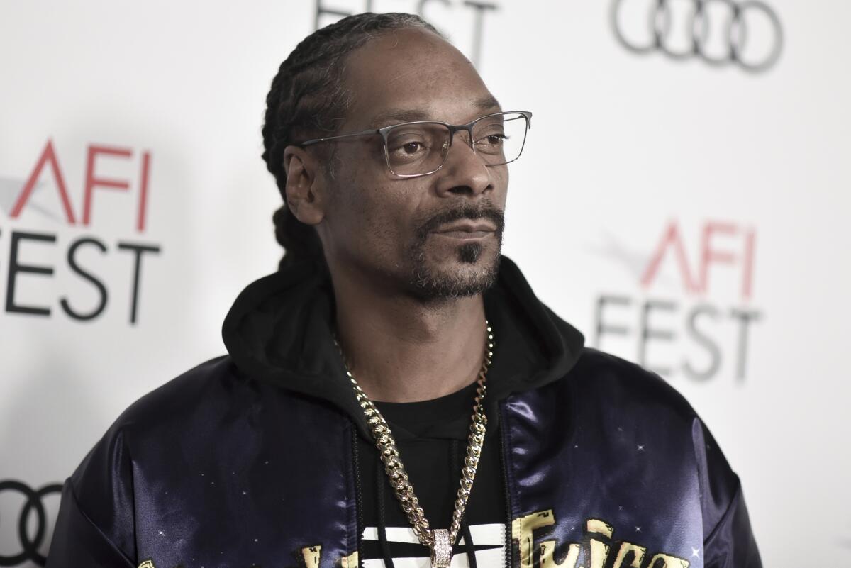 Snoop Dogg looks off to the side while wearing glasses, a black jacket and gold chain