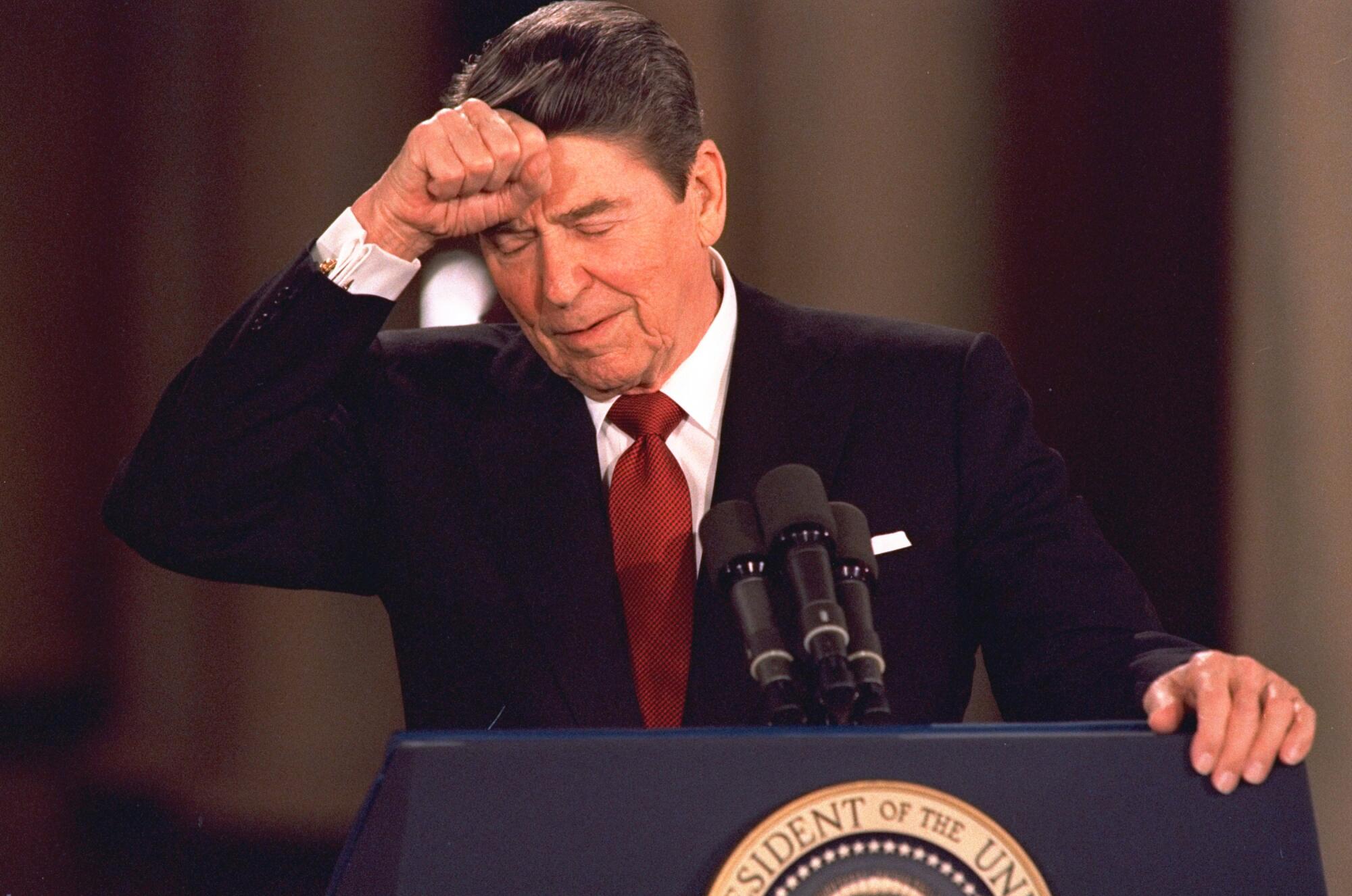 O presidente Reagan, de olhos fechados, mantém o punho direito na testa, a outra mão em um púlpito com o selo presidencial