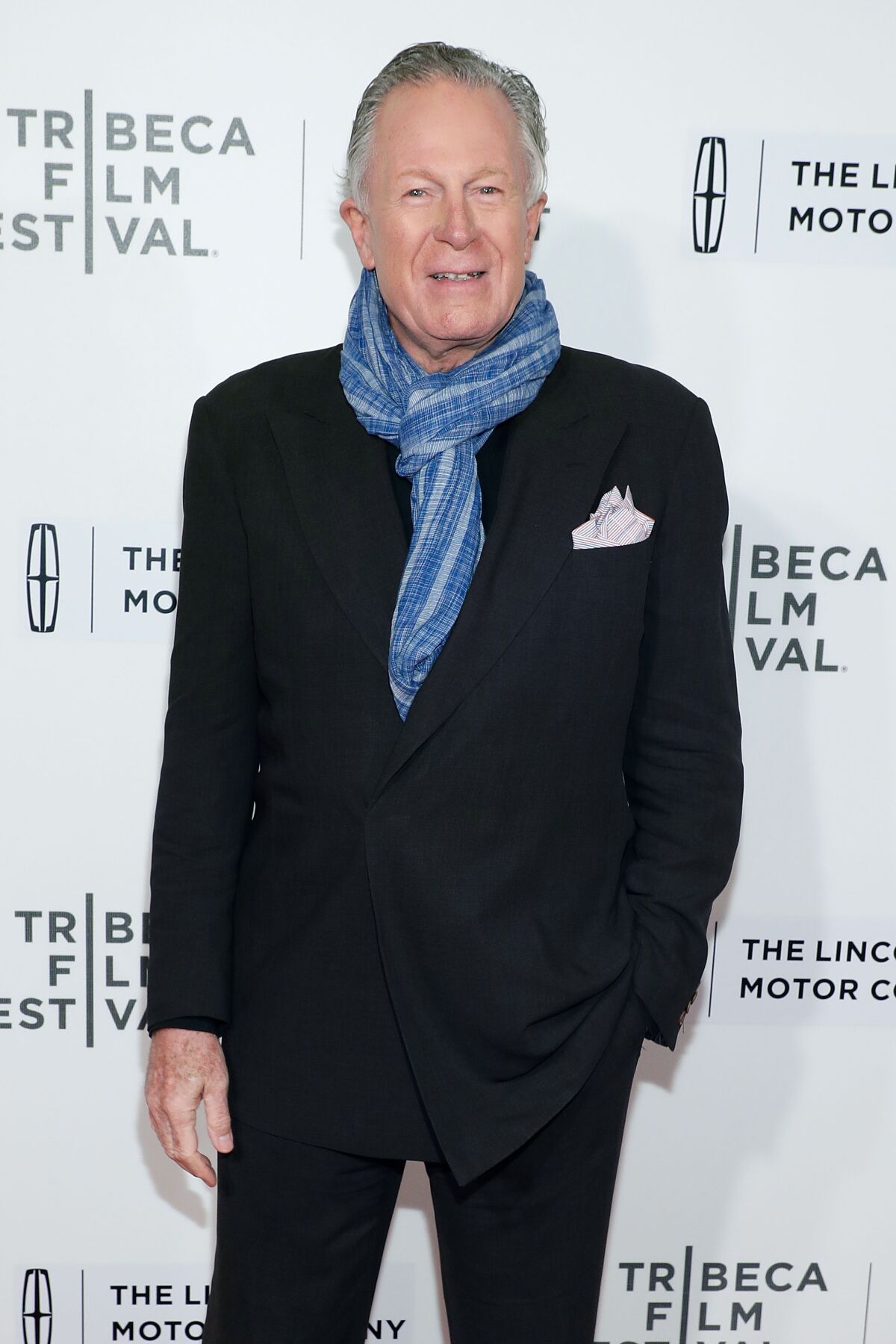 A man in a dark suit and blue scarf stands in front of a Tribeca Film Festival backdrop.