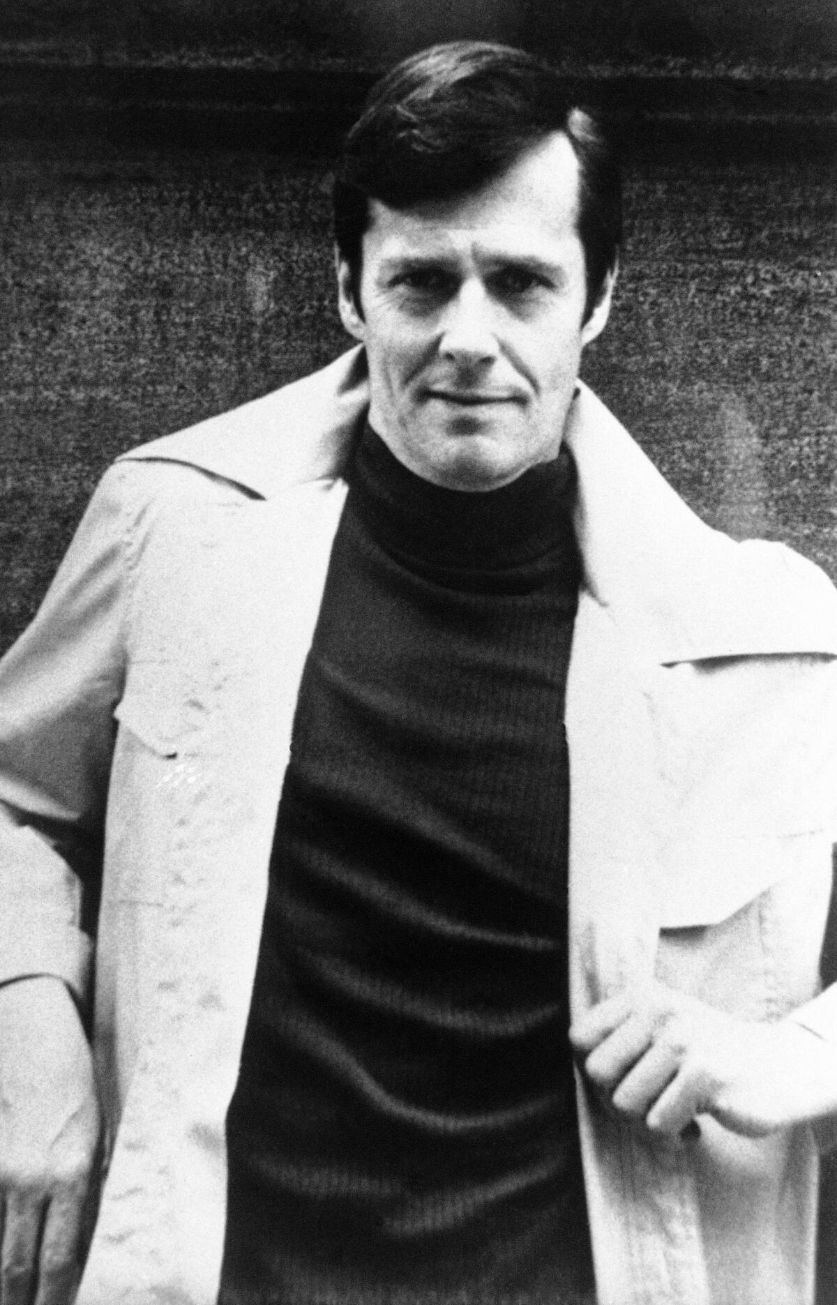 Ned Rorem, seen wearing a trench coat in a vintage black-and-white photo, stands with a hand at his waist.