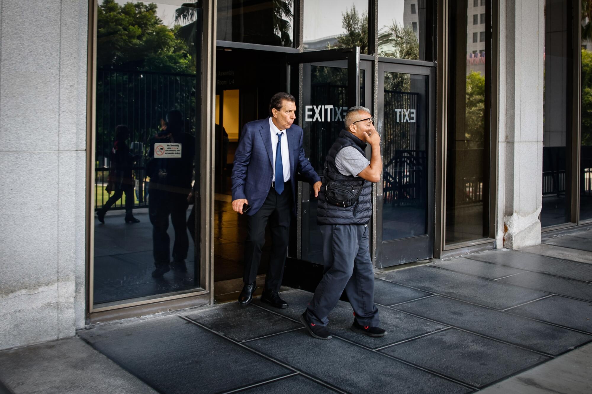 A man in a suit walks through the glass door of a courthouse behind another man.