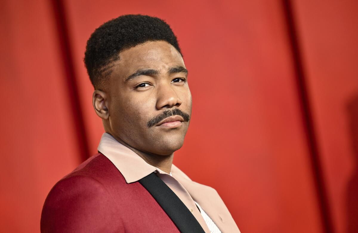 Donald Glover poses in a red suit in front of a red background.