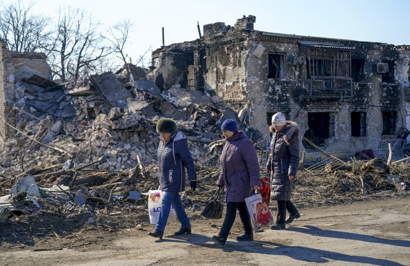Three women in warm hats and coats walk along a street lined with destroyed buildings: 