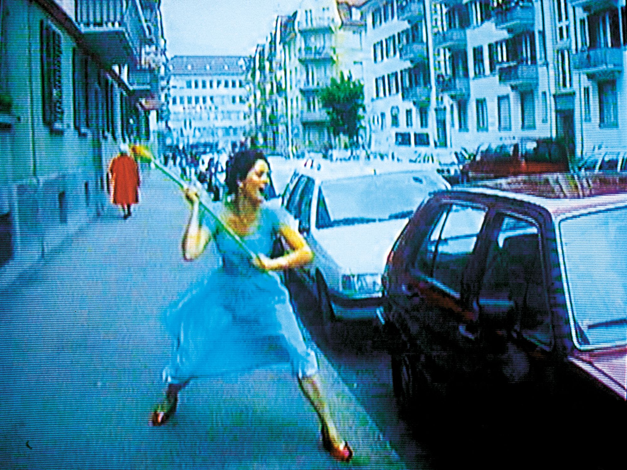 A woman in a sky blue dress and red shoes is seen on a sidewalk, about to smash a car window with a giant flower