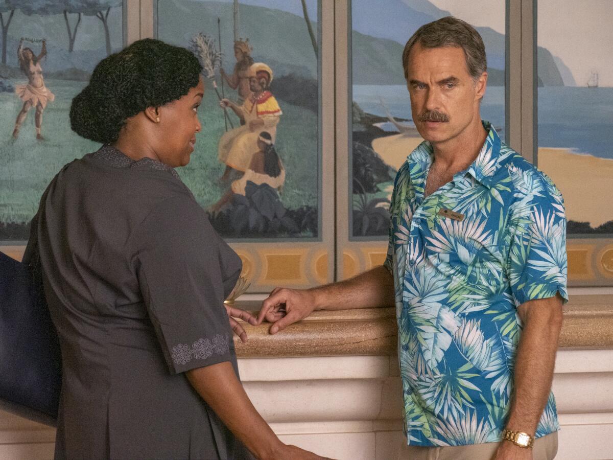 A man in a Hawaiian shirt and a woman in a black work uniform talk in front of a mural featuring native Hawaiians.